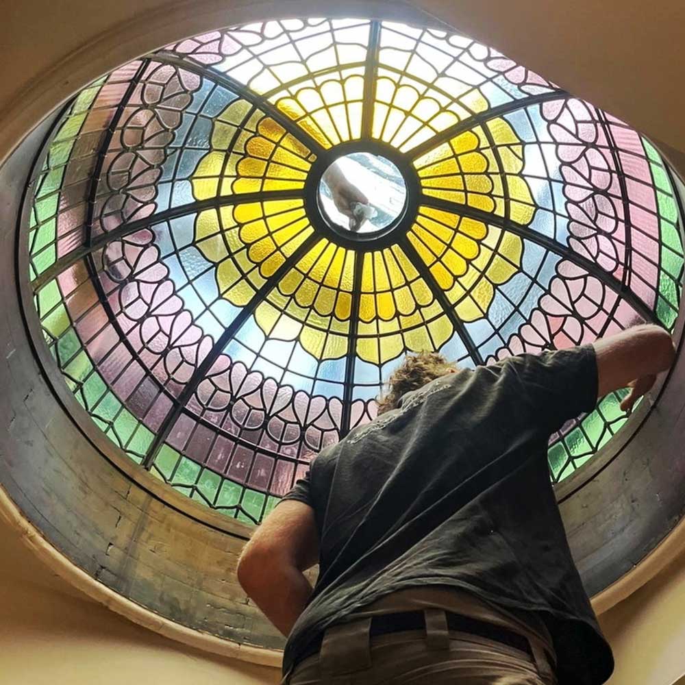 View from below of the newly reinstated stained glass dome at Rookery Hall Hotel, looking up through the restored lead glazing with a craftsman making final adjustments, showing the vivid yellows, pinks, blues and greens of the conserved glass panels