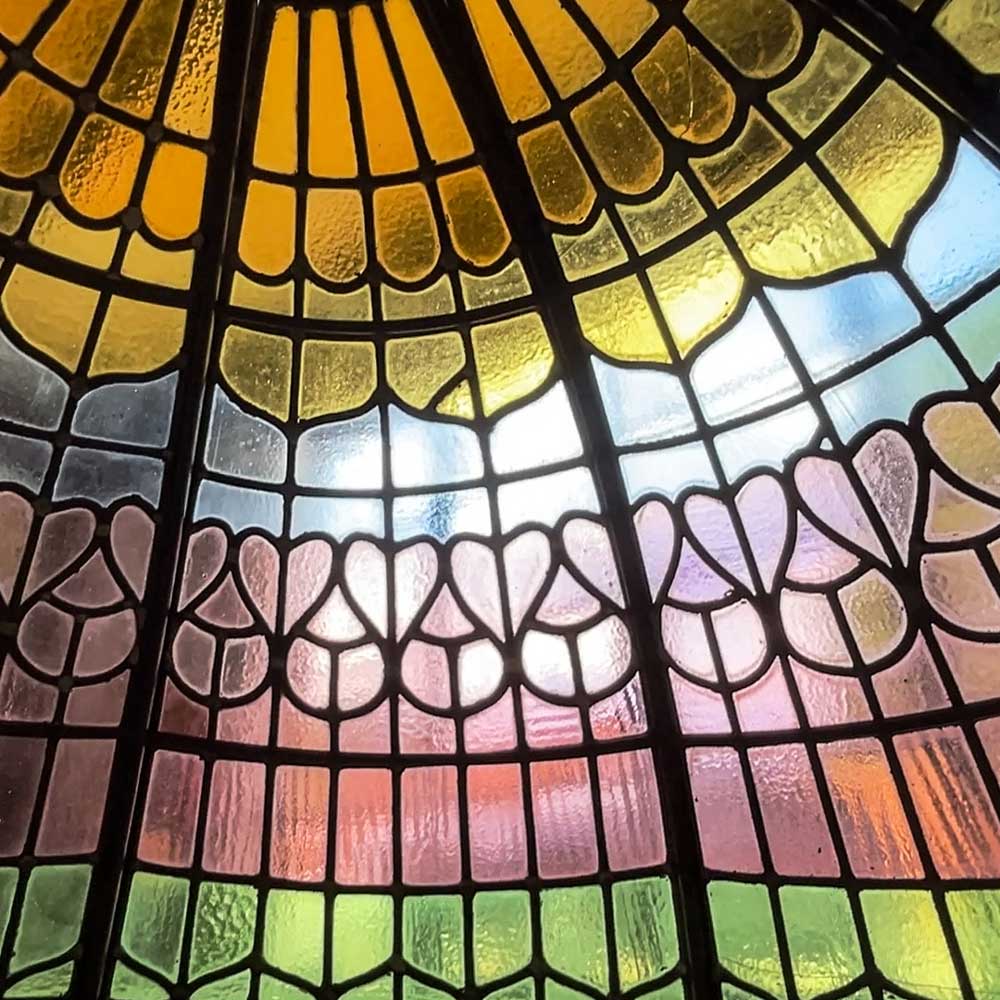 Interior close-up of the restored Victorian stained glass dome at Rookery Hall Hotel, showing the decorative scallop and petal motifs in amber, yellow, pale blue, mauve and green antique glass, backlit by natural daylight