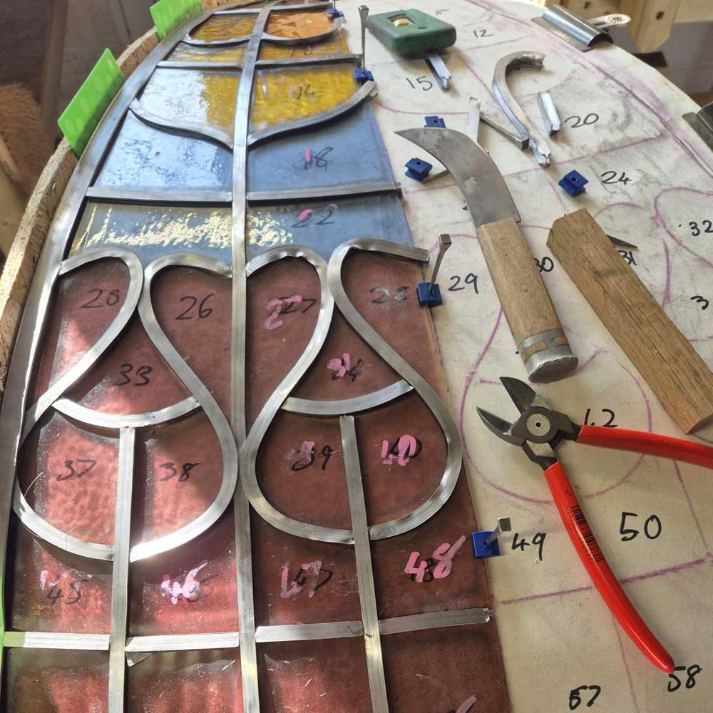 Restoration in progress on a curved dome panel in the Hawkscroft workshop, with restored lead came being fitted around numbered glass pieces against a full-size paper pattern, with specialist glazing tools visible alongside