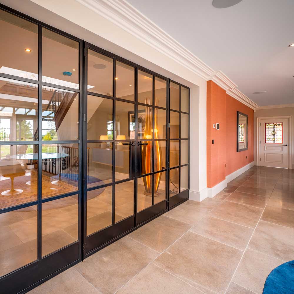 Internal hallway view of a large residential property featuring floor-to-ceiling black steel Crittal-style glazed doors and panels separating the corridor from an adjacent room
