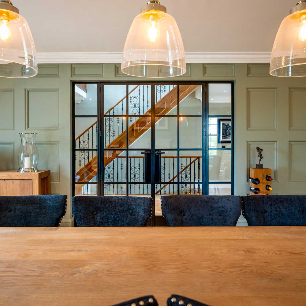 View from inside a formal dining room looking back through black metal glazed doors towards the entrance hall staircase