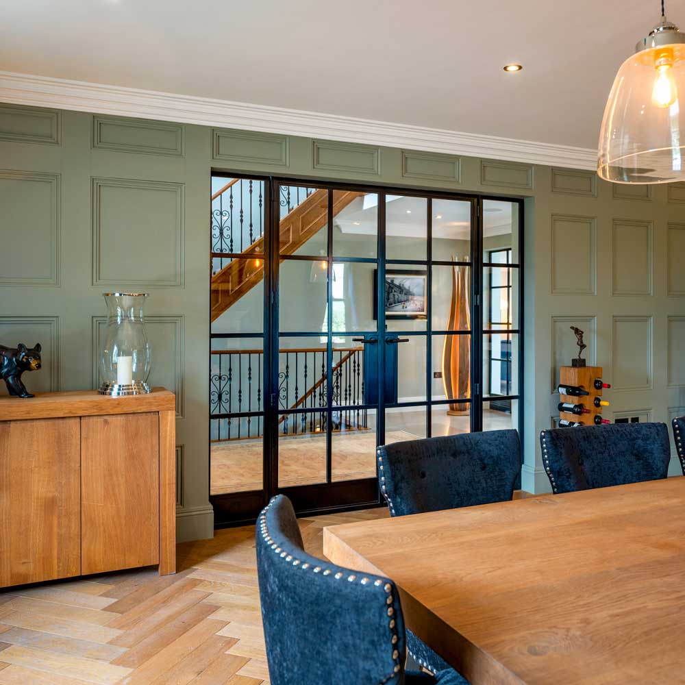 Interior of a formal dining room with panelled walls, viewed through partially open black steel Crittal-style glazed doors leading to the entrance hallway
