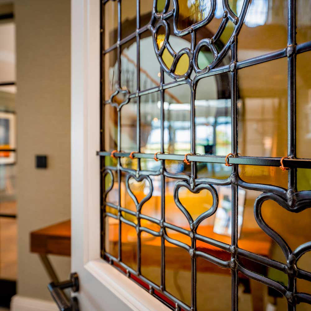 Close-up detail of a traditional leaded light stained glass window panel set into a white-painted door, showing heart-shaped and decorative lead came motifs with amber, green and clear glass panes