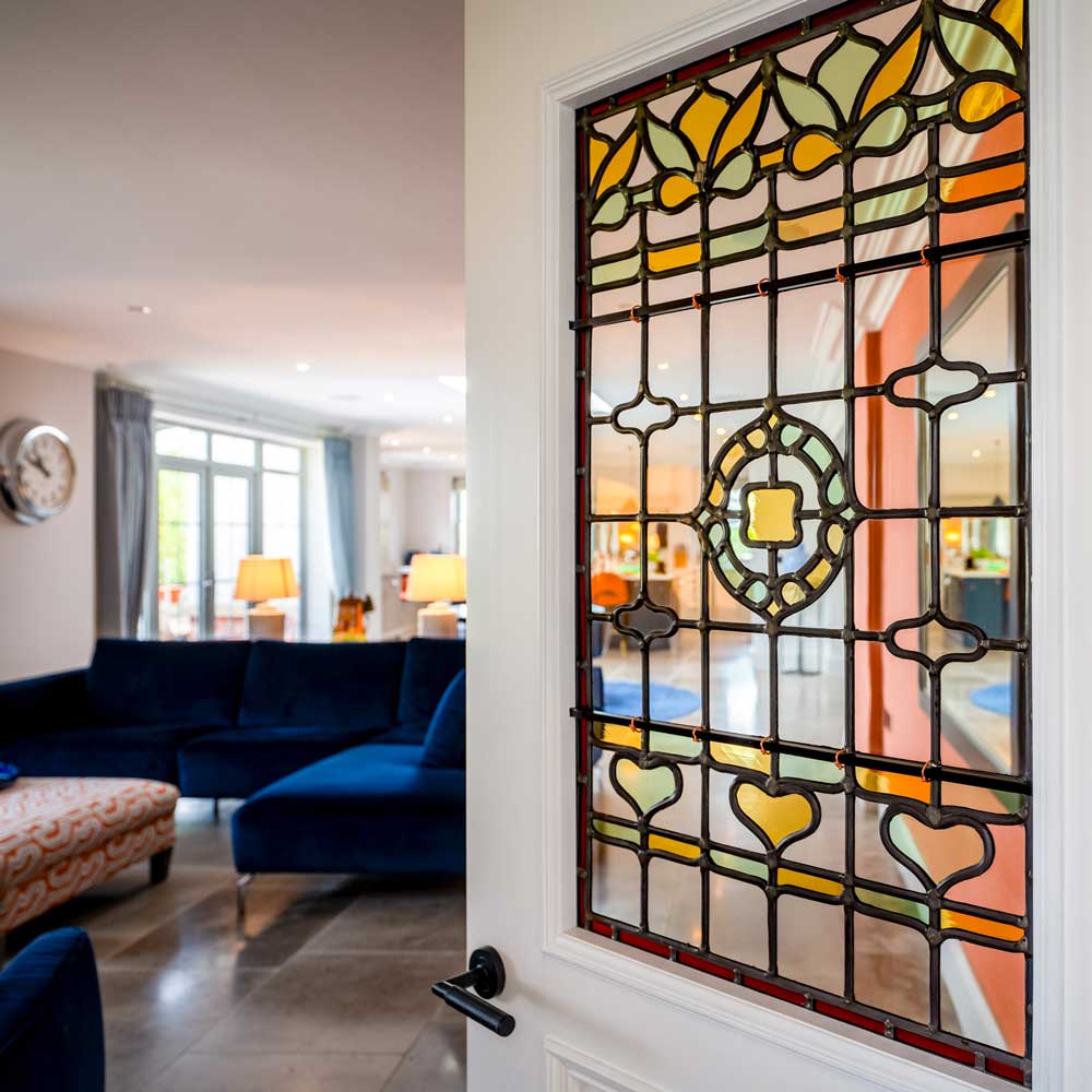 Traditional leaded light stained glass door panel with floral and geometric lead came designs in amber, green, orange and clear glass, set into a white-painted door with a black lever handle, looking through to a sitting room