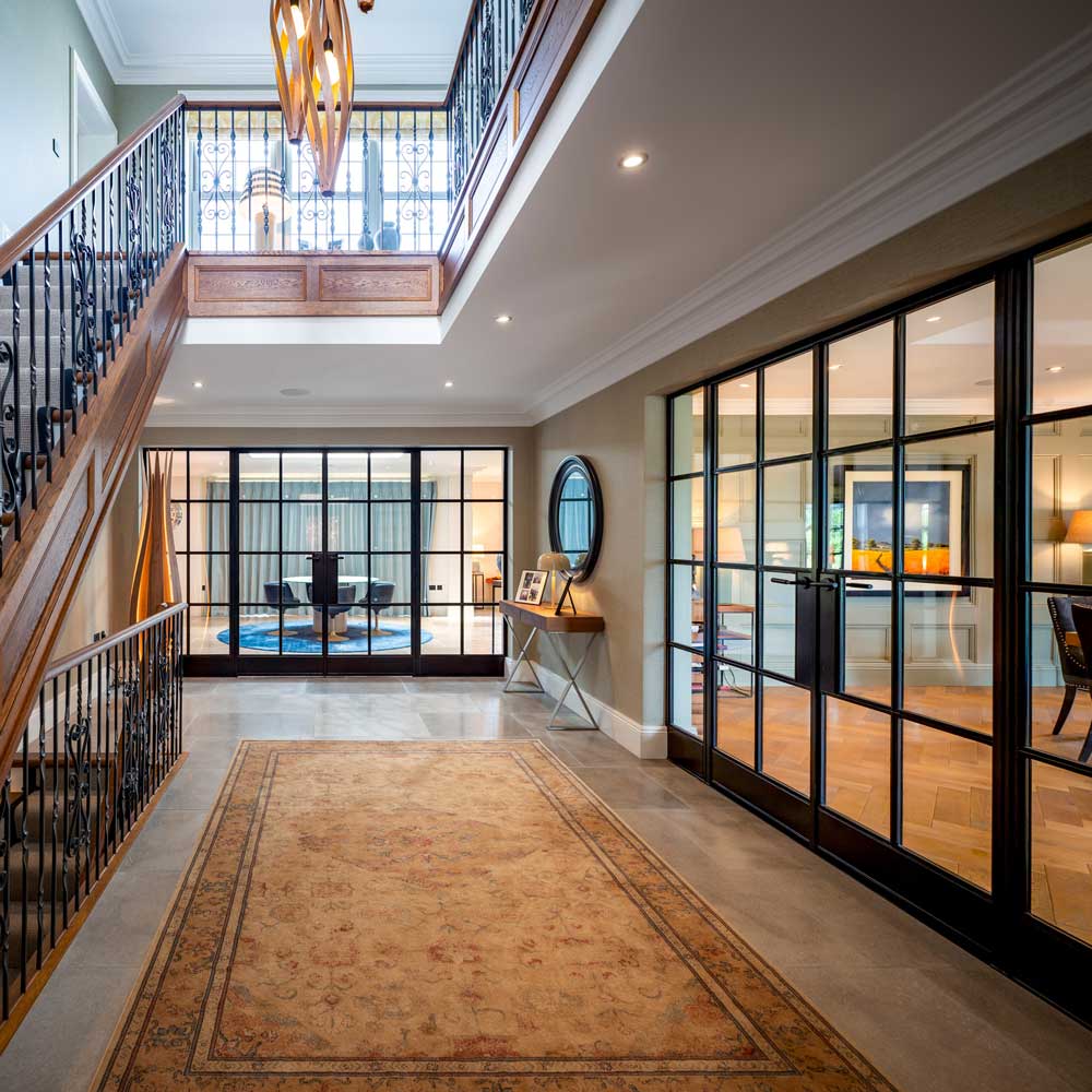 Wide-angle view of a grand double-height entrance hall. Multiple sets of black steel Crittal-style internal glazed doors opening to reception rooms on either side